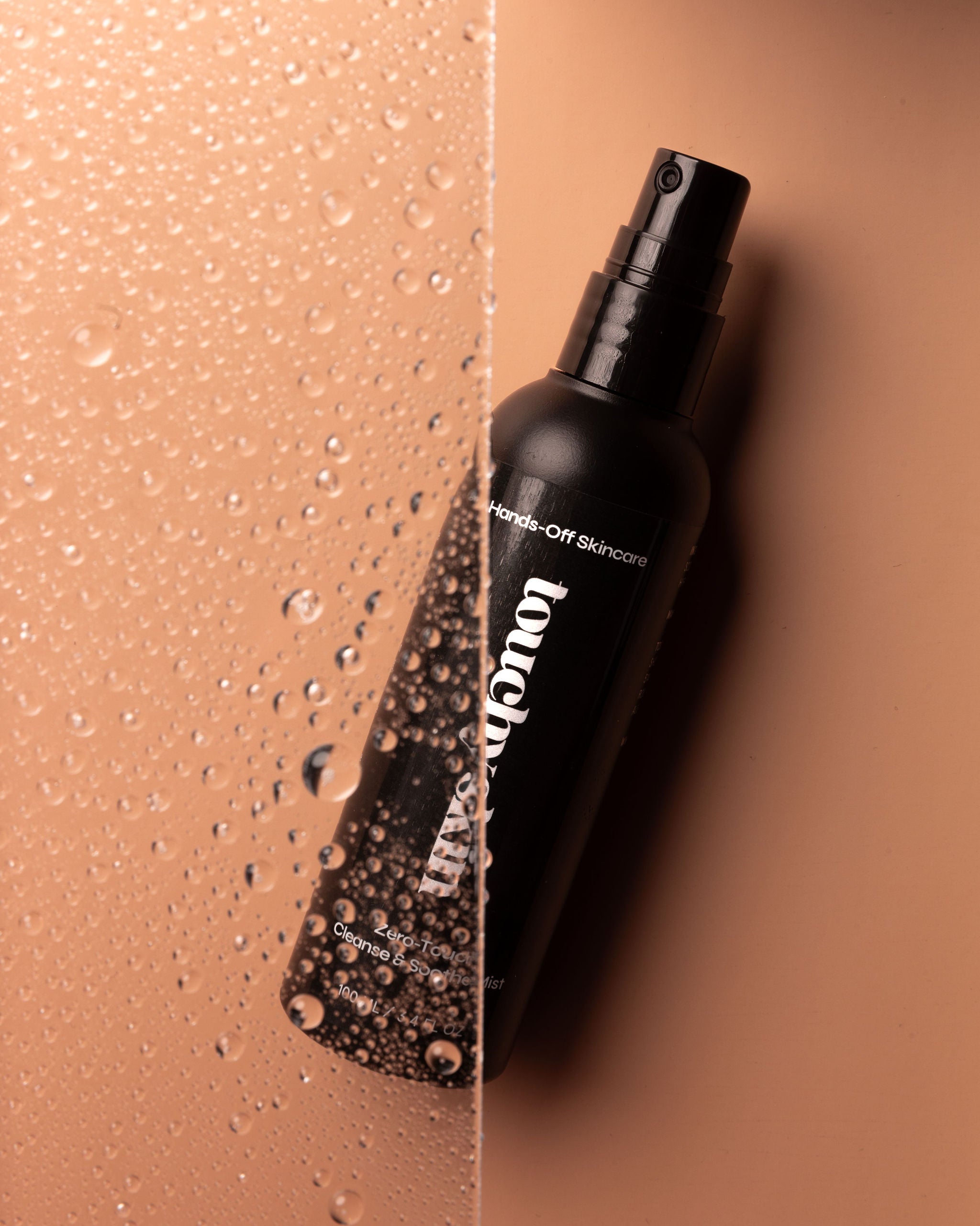 Touchy Skin Zero-Touch Cleanse & Soothe Mist bottle lying on a beige surface, partially obscured by a water-splashed glass panel.